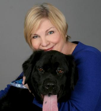 A smiling woman with short blonde hair in a blue top poses beside a big black dog with its long pink tongue out, against a gray background.